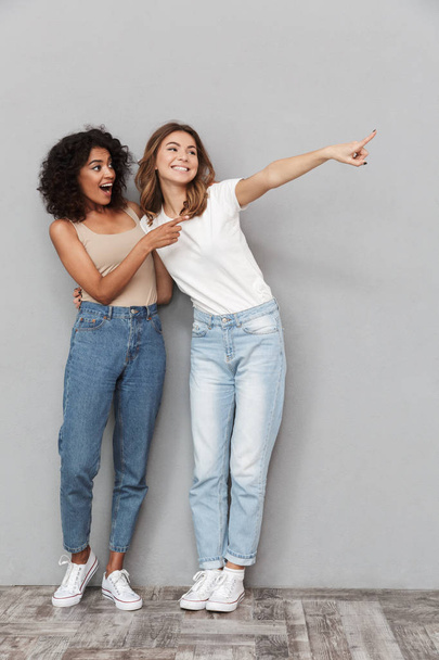 Full length portrait of two cheerful young women standing together and pointing away at copy space over gray background - Fotoğraf, Görsel