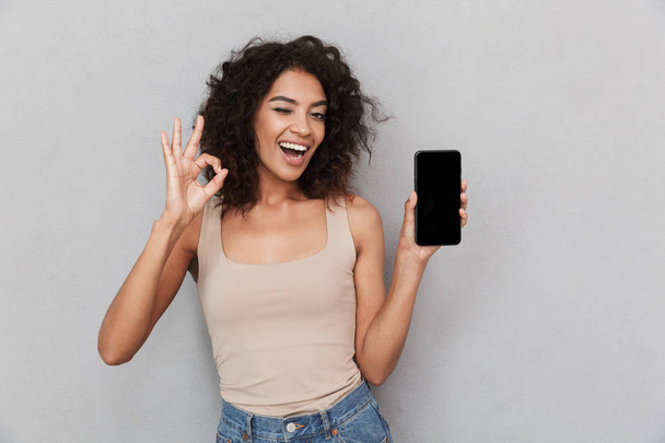 Portrait of a smiling young african woman holding blank screen mobile phone and showing ok over gray background - Φωτογραφία, εικόνα