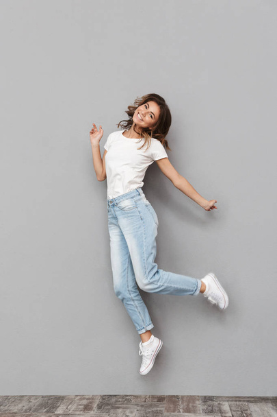 Full length portrait of a happy young woman jumping and celebrating over gray background - Fotografie, Obrázek