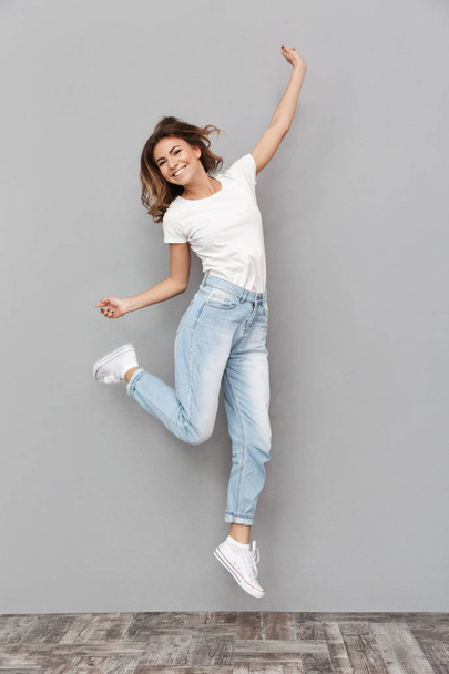 Full length portrait of a cheerful young woman jumping and celebrating over gray background - 写真・画像