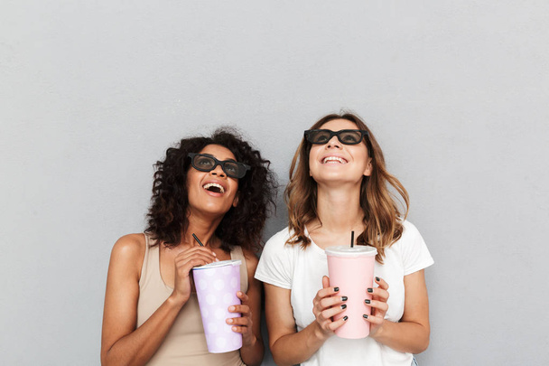 Portrait of two smiling young women in 3d glasses holding plastic cups and looking up isolated over gray background - Фото, зображення