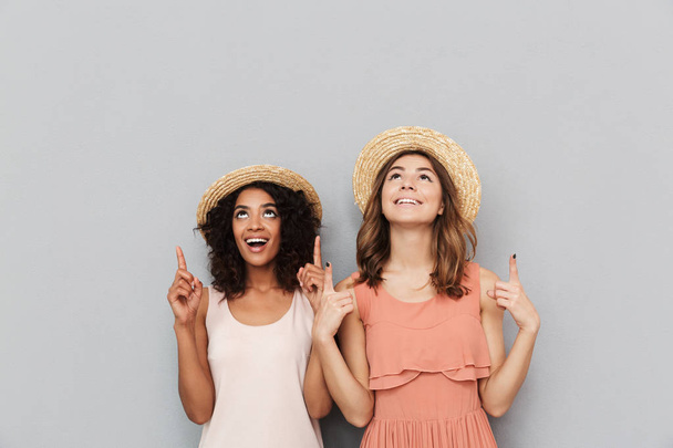 Портрет двох щасливих багатоетнічних жінок, кавказьких і афроамериканських дівчат у солом'яних капелюхах, посміхаючись, вказуючи пальці вгору на копійку ізольовано на сірому фоні
 - Фото, зображення
