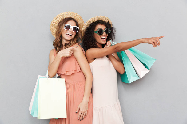 Portrait of two lovely young women dressed in summer clothes holding shopping bags and pointing at copy space isolated over gray background - Foto, imagen