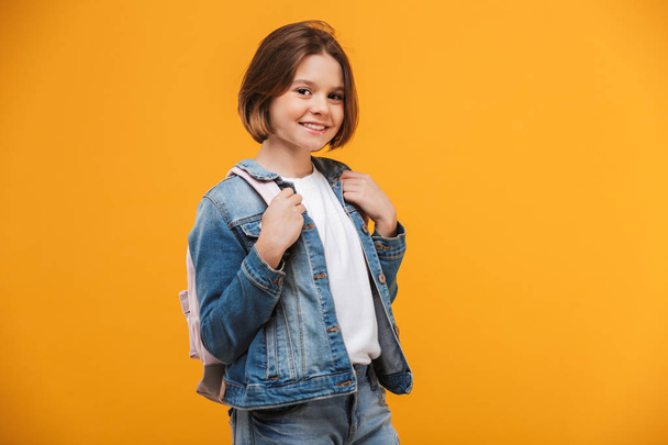 Portrait of a smiling little schoolgirl with backpack looking at camera over yellow background - Valokuva, kuva