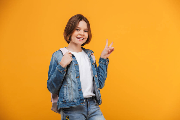 Portrait of a smiling little schoolgirl with backpack looking at camera and showing peace gesture over yellow background - Fotoğraf, Görsel