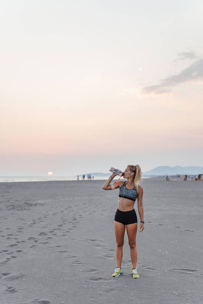 Hübsche Sportlerin steht am Sandstrand und trinkt Wasser. - Foto, Bild