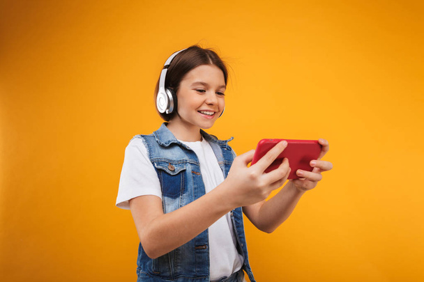 Portrait of a satisfied little schoolgirl listening to music with headphones while using mobile phone over yellow background - Photo, Image