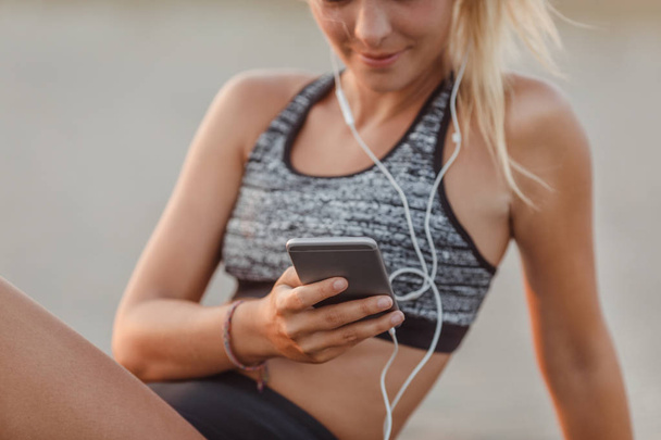 Onherkenbare Sportvrouw zittend op een zandstrand en het houden van haar mobiele telefoon. - Foto, afbeelding
