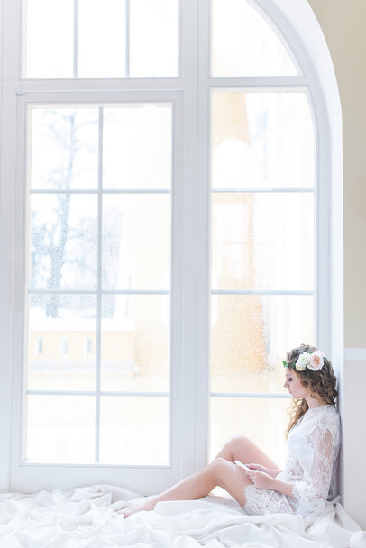 Charming young curly bride in white sitting on a huge light window - Foto, Imagem