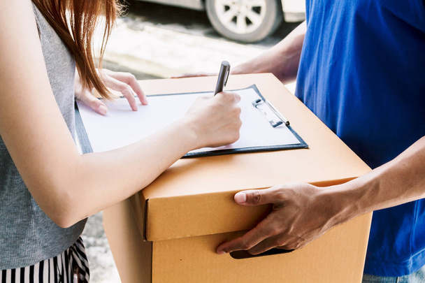 Woman putting signature in clipboard on cardboard box with delivery man  - Foto, afbeelding