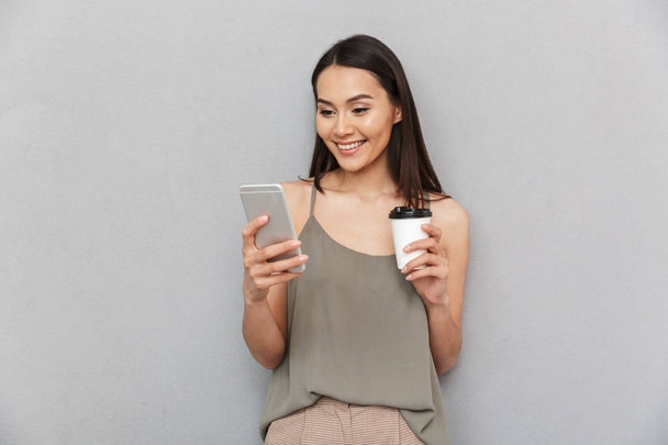 Portrait of a smiling asian woman holding takeaway coffee cup and using mobile phone isolated over gray background - Fotó, kép