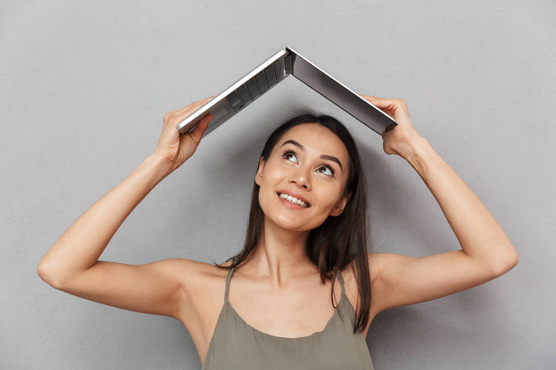Portrait of a smiling asian woman holding laptop computer above her head isolated over gray background - Valokuva, kuva