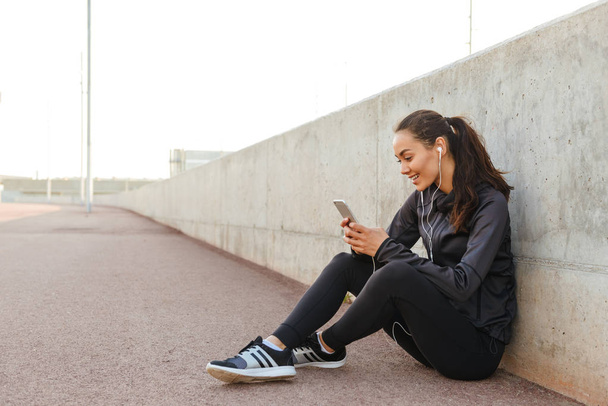 Photo of cheerful young asian sports woman sitting outdoors listening music using mobile phone. - Foto, Imagen