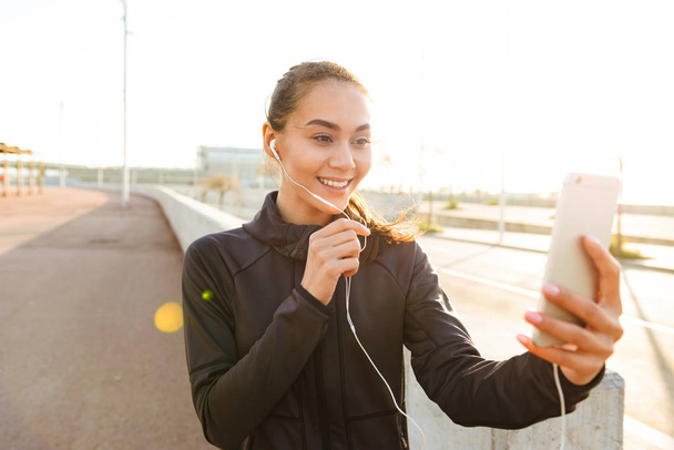 Image of happy young asian sports woman walking outdoors talking by mobile phone. - Fotoğraf, Görsel