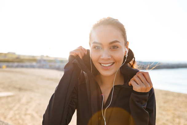 Photo of cheerful young asian sports woman sitting outdoors listening music looking camera. - Foto, Imagem