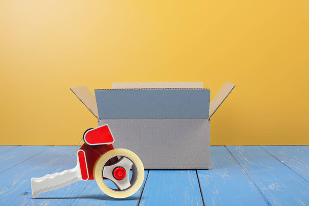 Poštovné a balné služby - otevřený balíček a pásky dávkovač čelní pohled na pozadí modré dřevo a žluté zdi. - Fotografie, Obrázek