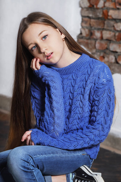 Portrait stylish model child. Beautiful teenager in blue sweater. Fashion pose model. Young hipster girl sitting chair, looking at camera confident, defiant. Studio shot. hands on face. - Valokuva, kuva