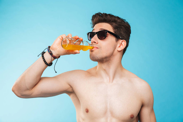 Close up portrait of a shirtless young man in sunglasses drinking beer from a bottle over blue background - Foto, Bild
