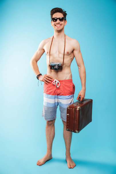 Full length portrait of a cheerful young shirtless man in swimming shorts carrying photo camera and suitcase over blue background - 写真・画像