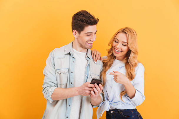 Photo of happy people man and woman laughing while pointing finger at smartphone isolated over yellow background - Valokuva, kuva