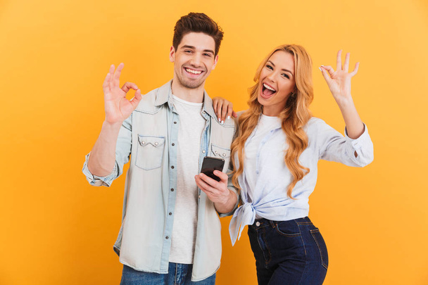Photo of joyous couple man and woman smiling and showing ok signs while holding smartphone isolated over yellow background - Φωτογραφία, εικόνα