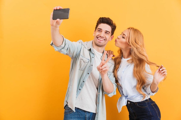 Portrait of lovely couple taking selfie photo on cell phone while woman kissing man on his cheek isolated over yellow background - 写真・画像