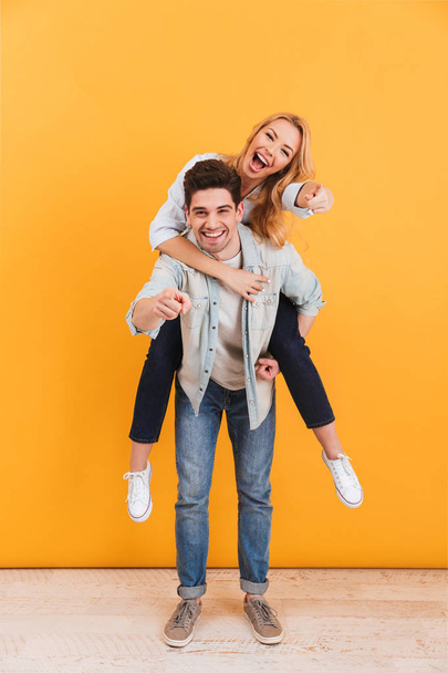 Full length photo of smiling couple having fun and pointing fingers at you while man piggybacking happy woman isolated over yellow background - Zdjęcie, obraz