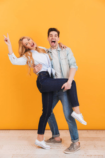 Full length photo of young couple having fun and hugging at camera while happy woman showing peace sign isolated over yellow background - Foto, Bild