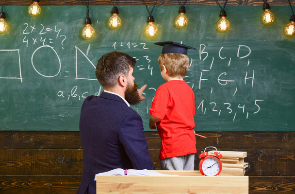 先生の助けを借りて、方程式を解く卒業キャップの生徒。男は数字でボードを指さしながら小さな子供の算数を指導します。成人男性と少年が教室で引き返した. - 写真・画像