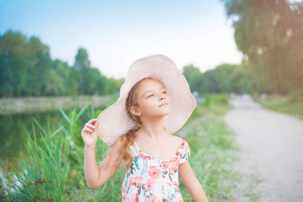 Kaunis koulutyttö vaaleanpunaisessa hatussa auringonlaskun säteiltä kesäiltana luonnossa kaupungin ulkopuolella.
. - Valokuva, kuva