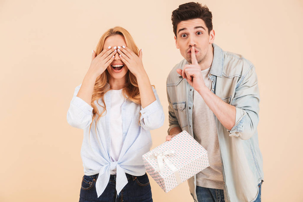 Image of brunette man with present box keeping finger at lips and showing shh while standing near woman covering eyes with hands isolated over beige background - Foto, Imagen
