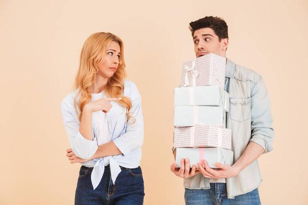 Portrait of disappointed woman pointing finger at upset man standing with lots of gift boxes isolated over beige background - Foto, immagini