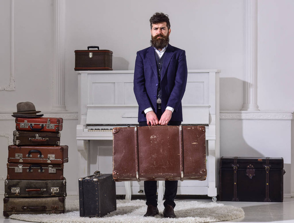 Baggage and travelling concept. Man, traveller with beard and mustache with baggage, luxury white interior background. Macho stylish on strict face stands and carries big vintage suitcase. - Фото, зображення