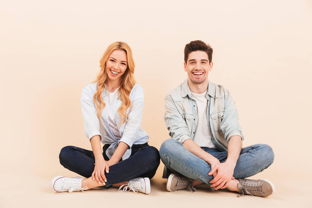 Image of attractive joyful couple man and woman 20s in denim clothing smiling while sitting on the floor with legs crossed isolated over beige background - Fotó, kép