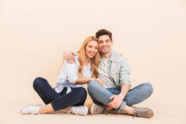 Image of nice and cute couple in denim clothing smiling and hugging together while sitting on the floor with legs crossed isolated over beige background - Fotografie, Obrázek