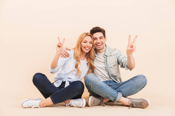 Image of pleased man and woman hugging together while sitting on the floor with legs crossed and showing peace symbols isolated over beige background - Foto, Imagem