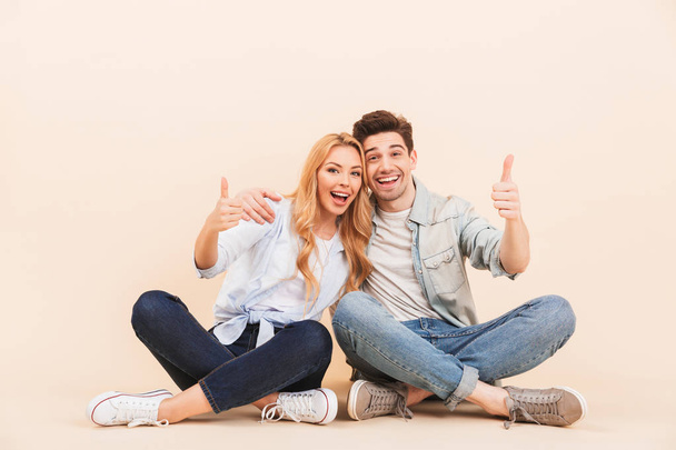 Image of handsome man and beautiful woman hugging together while sitting on the floor with legs crossed and showing thumbs up isolated over beige background - Foto, afbeelding