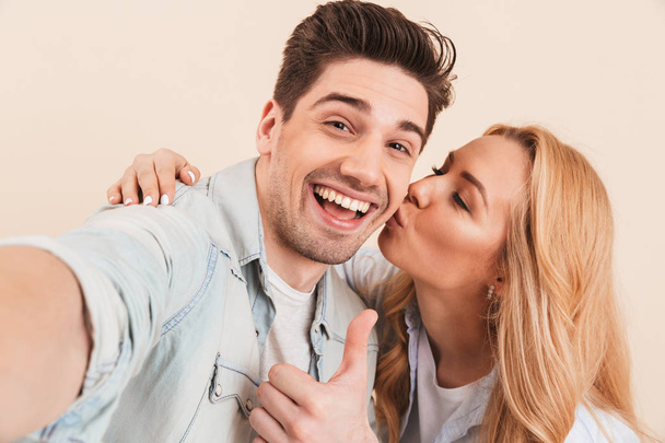 Portrait of happy man taking selfie photo and gesturing thumb up at camera while adorable woman kissing his cheek isolated over yellow background - Foto, imagen