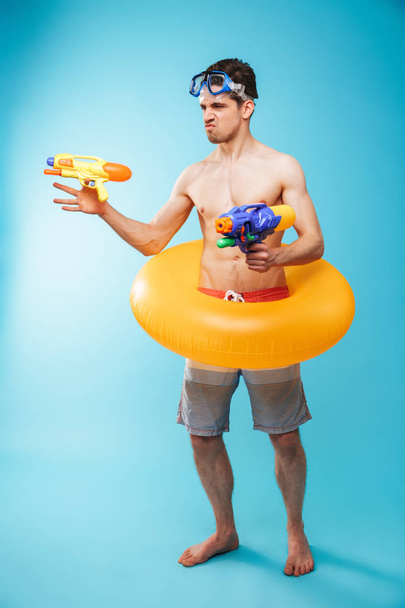 Full length portrait of an excited young shirtless man in swim goggles and inflatable ring having fun with water guns over blue background - Фото, зображення