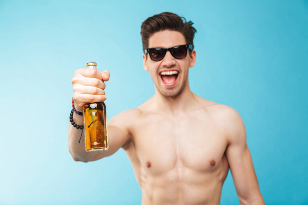 Portrait if a cheerful shirtless man showing beer bottle over blue background - Фото, изображение