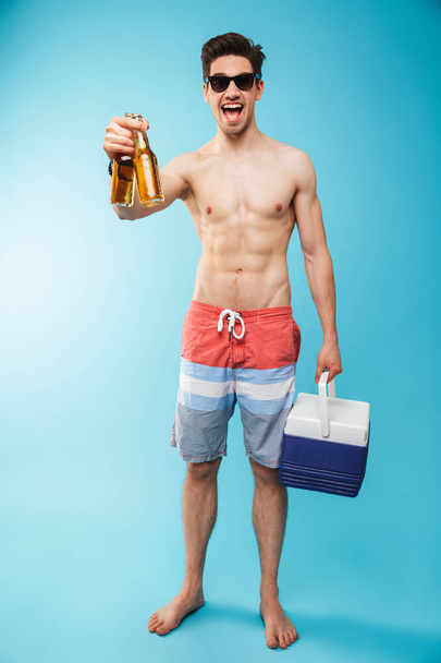 Full length portrait if an excited shirtless man in swimming shorts holding cooler bag and beer bottles over blue background - Photo, image