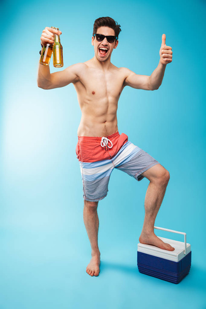Full length portrait if a cheerful shirtless man in swimming shorts holding beer bottles and showing thumbs up over blue background - Zdjęcie, obraz