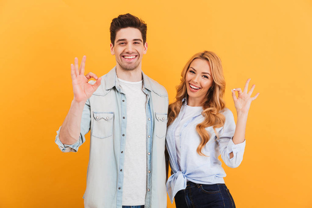 Photo of optimistic people man and woman in basic clothing smiling and gesturing ok symbol at camera isolated over yellow background - Photo, Image