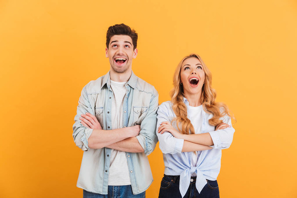 Image of excited people man and woman in basic clothing posing with arms crossed and looking upward isolated over yellow background - 写真・画像