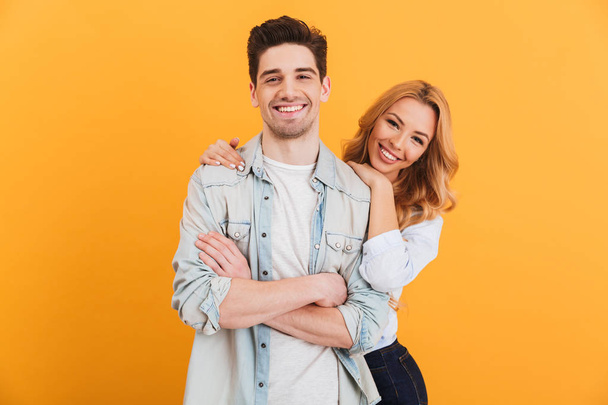Portrait of loving couple man and woman in basic clothing looking at camera with happy smile isolated over yellow background - 写真・画像