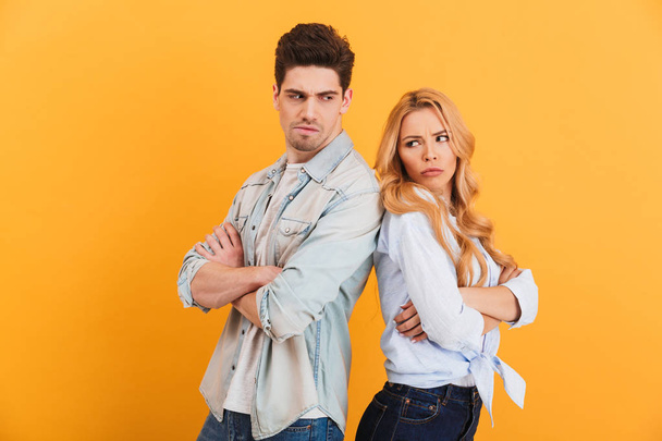 Photo of resented man and woman in denim clothes standing back to back with arms folded in quarrel isolated over yellow background - Fotografie, Obrázek