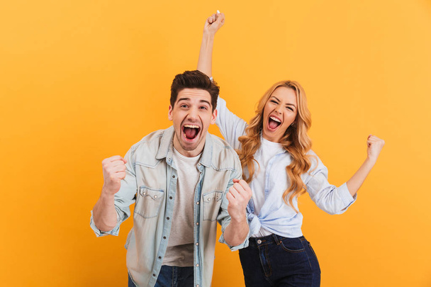 Portrait of cheerful people man and woman in basic clothing smiling and clenching fists like winners or happy people isolated over yellow background - Photo, image
