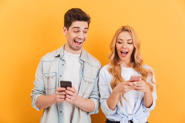 Image of funny young couple man and woman peeking at each others cell phones holding in hands isolated over yellow background - Foto, imagen