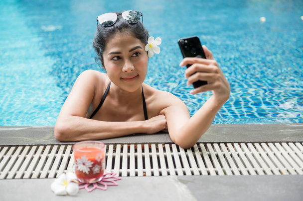 Attractive sexy Asian woman with black two piece swimsuit and sunglasses selfie herself by smartphone nearat swimming pool edge. Beautiful Girl relax during summer vacation. - Photo, Image