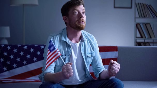 Emotional American man holding USA flag and watching election results on tv home - 写真・画像